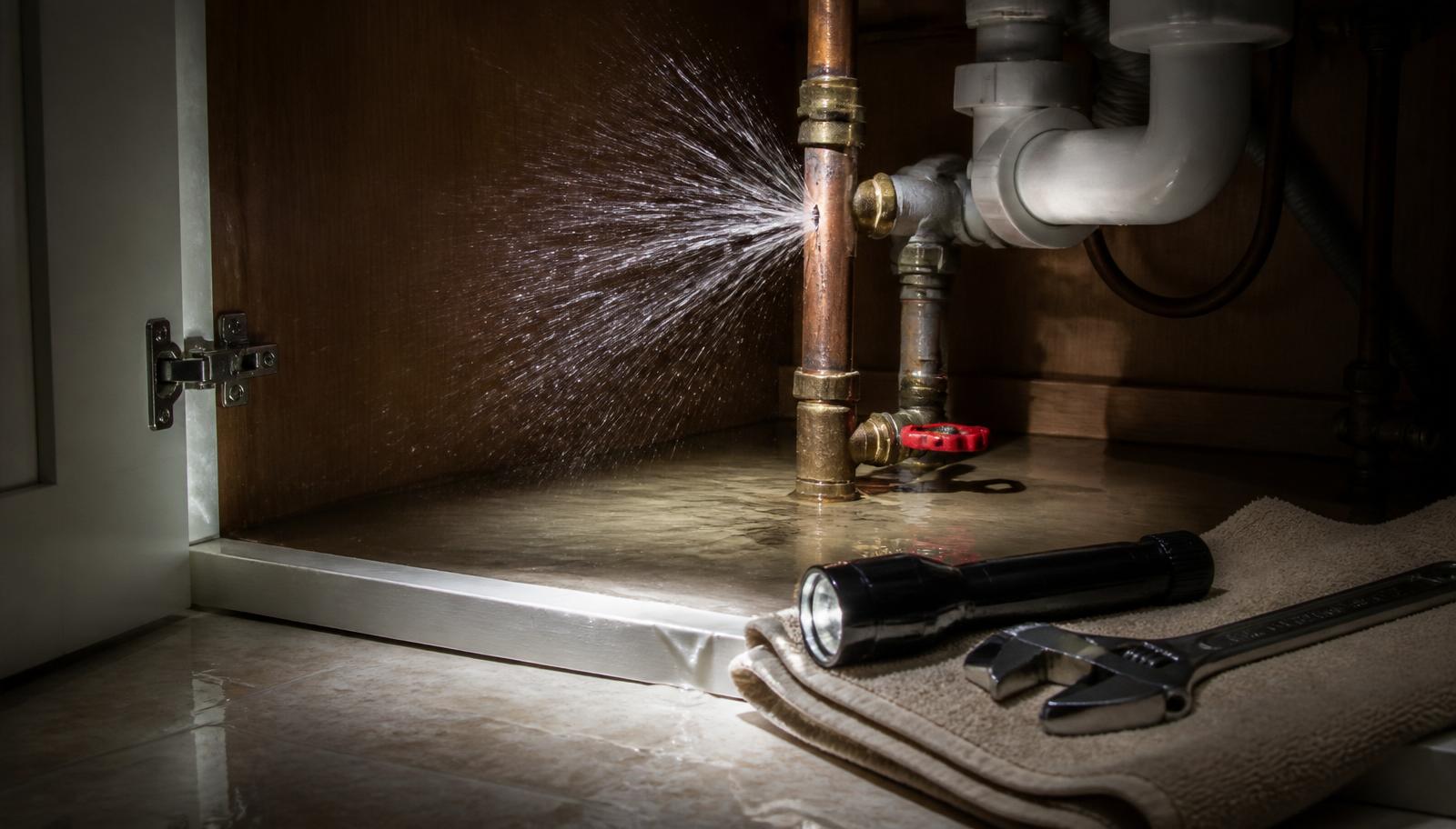 Burst copper water pipe spraying under a kitchen sink during an emergency plumbing call-out